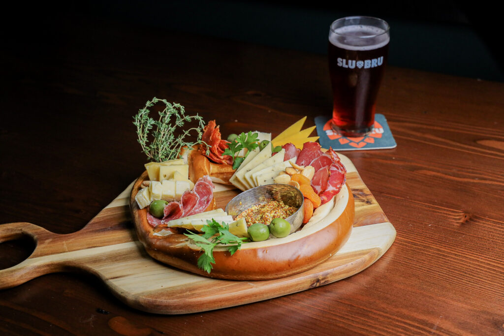 A wooden cutting board displays a plate of Seattle’s Best Bavarian Pretzel Combo with cured meats, cheese, olives, and mustard.